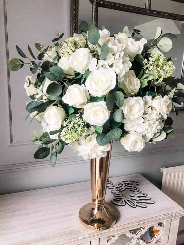A gold vase filled with white roses and greenery, perfect for decoration hire.