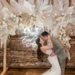 A bride and groom kissing in front of a wedding arch with feathers.